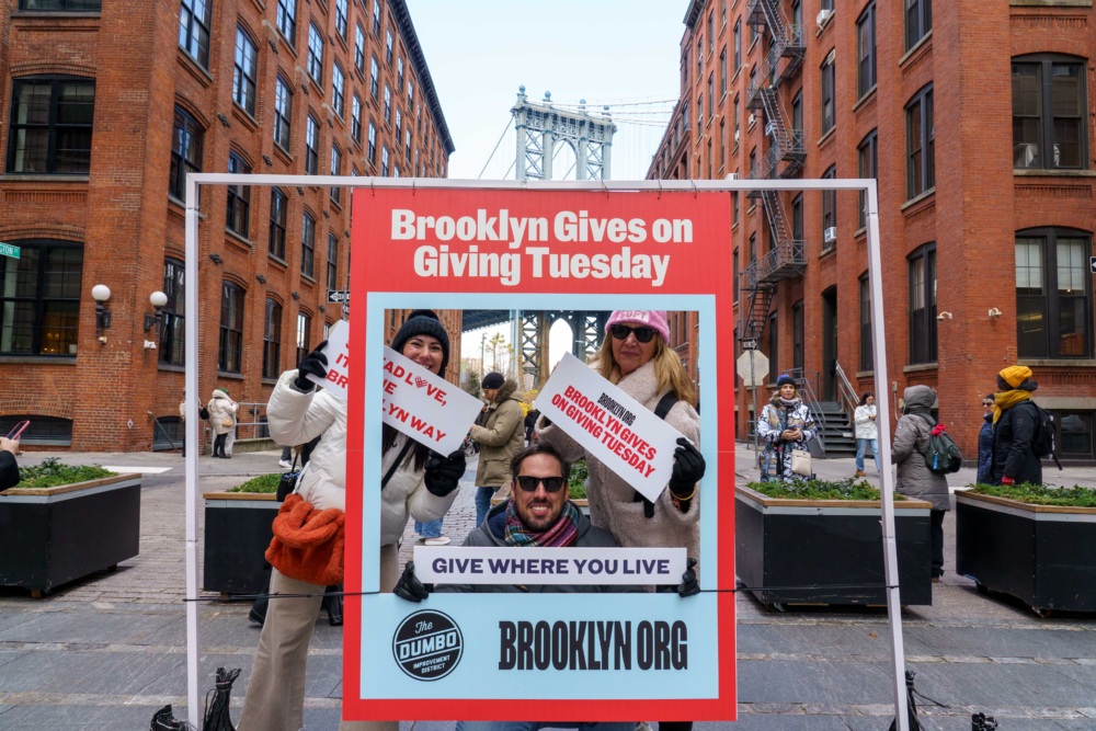 People posing inside a large picture frame with "Brooklyn Gives on Giving Tuesday" text, in front of the Manhattan Bridge, amid brick buildings and a busy street.