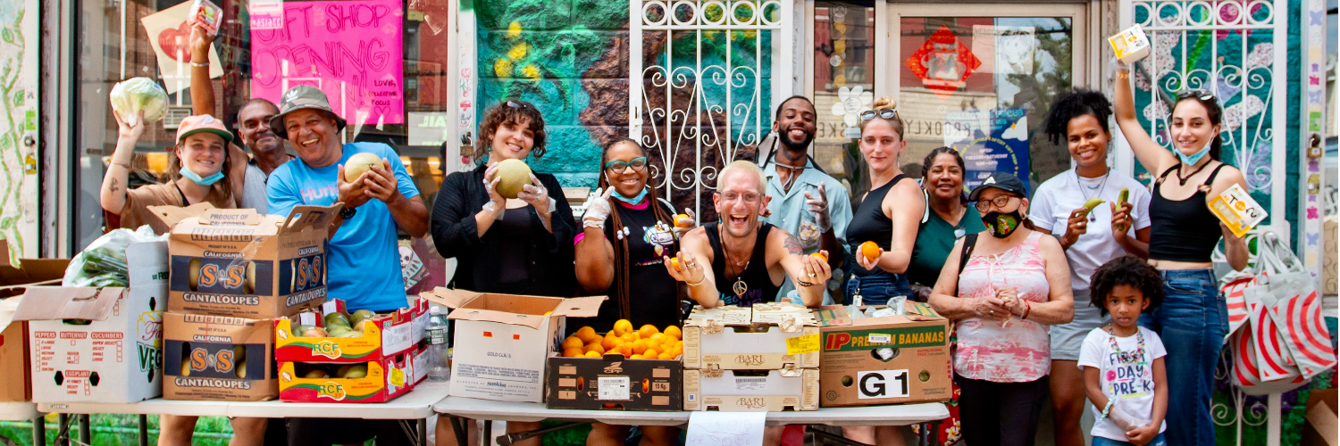 A diverse group of people standing behind tables filled with various fruits and vegetables in front of a storefront. Some people are smiling and holding produce.