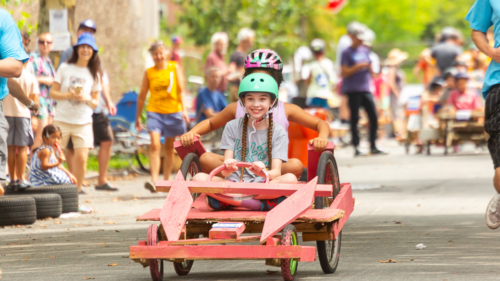 Two children wearing helmets are riding a pink handmade go-kart in a race. Spectators line the street, watching and cheering. The event takes place outdoors on a sunny day.