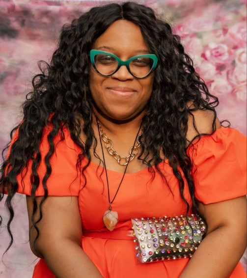 A person with long curly hair, wearing green glasses and an orange off-the-shoulder dress, holds a studded clutch purse. The background features a soft pink and purple floral pattern.