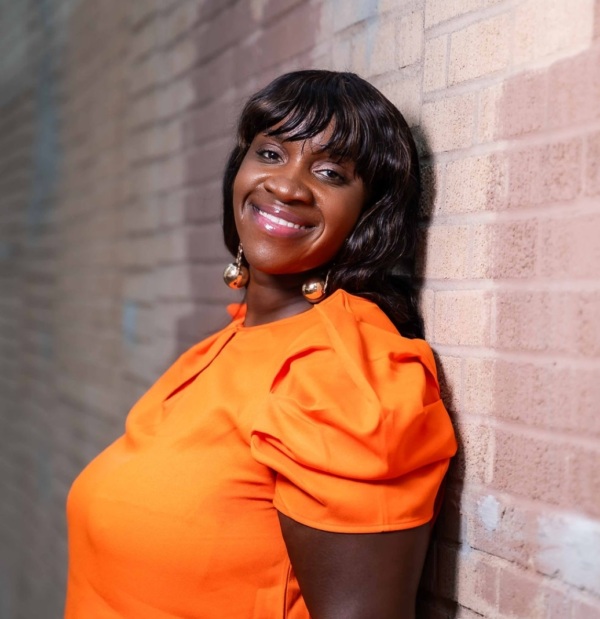 A woman wearing an orange blouse and earrings, smiling and leaning against a brick wall.