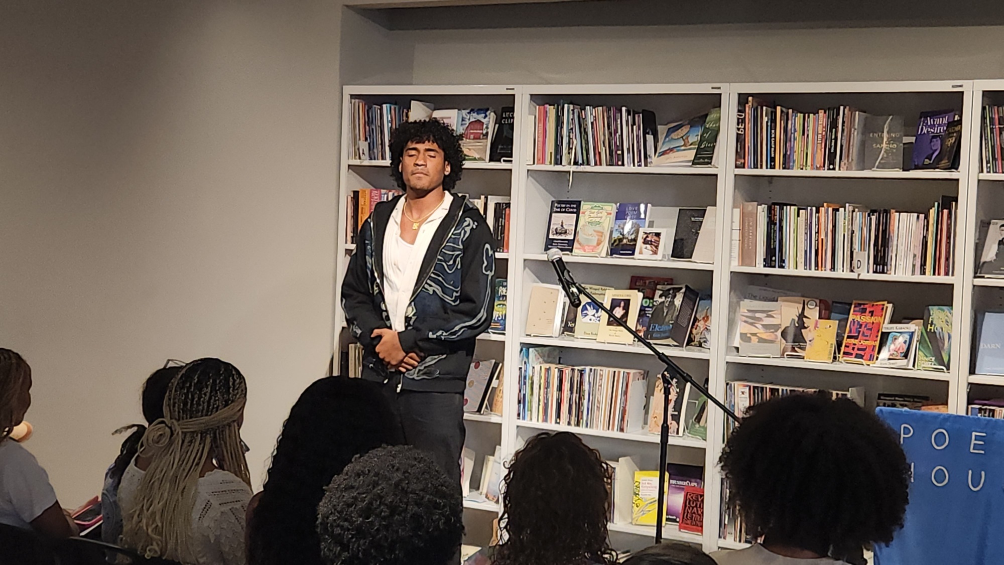 A person stands in front of a bookshelf, speaking or performing to an audience seated in a library or bookstore. A microphone stand is in front of them.