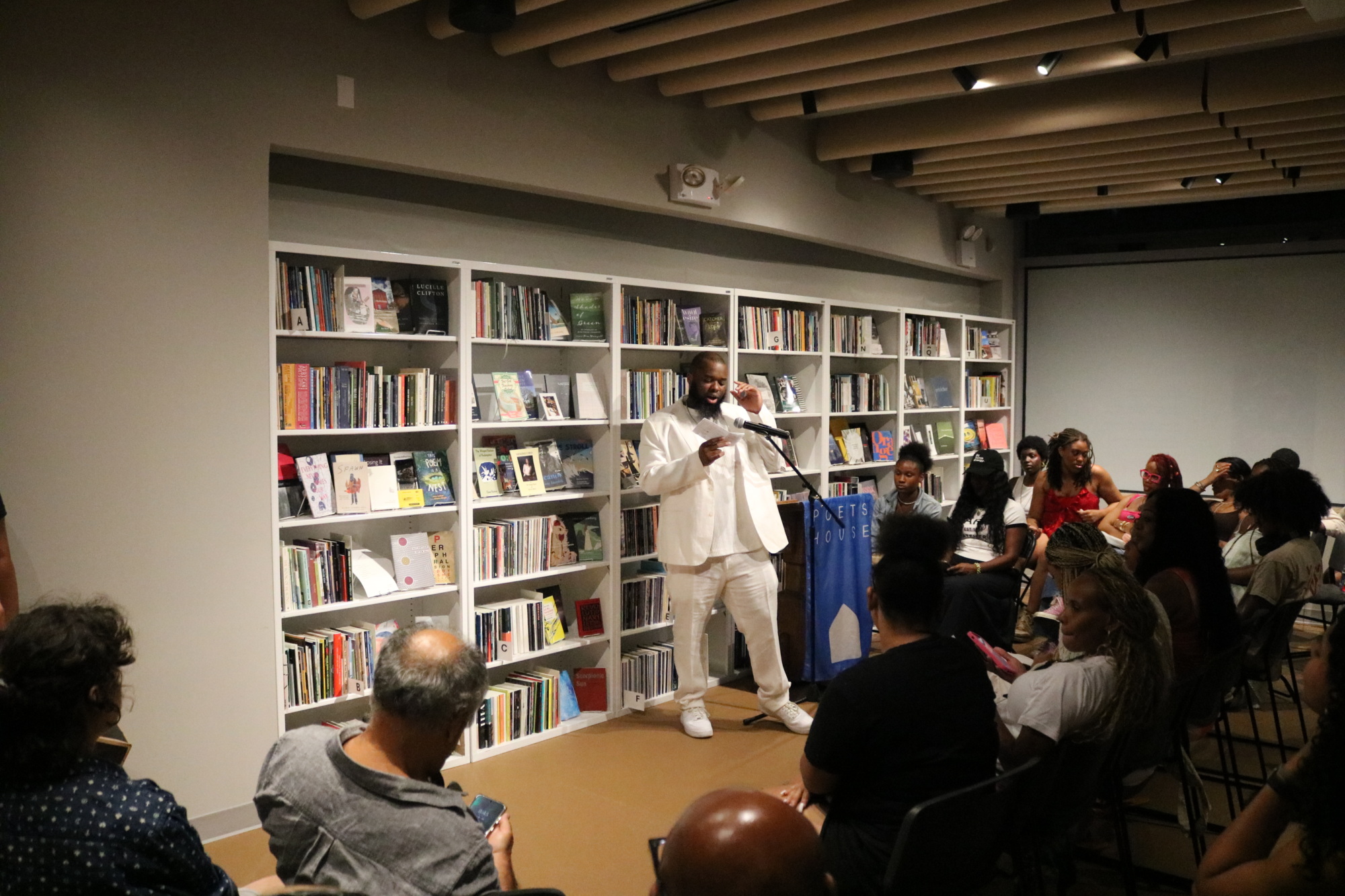 A man in white speaking at a podium in front of an audience at a bookstore event. Shelves of books line the wall behind him.