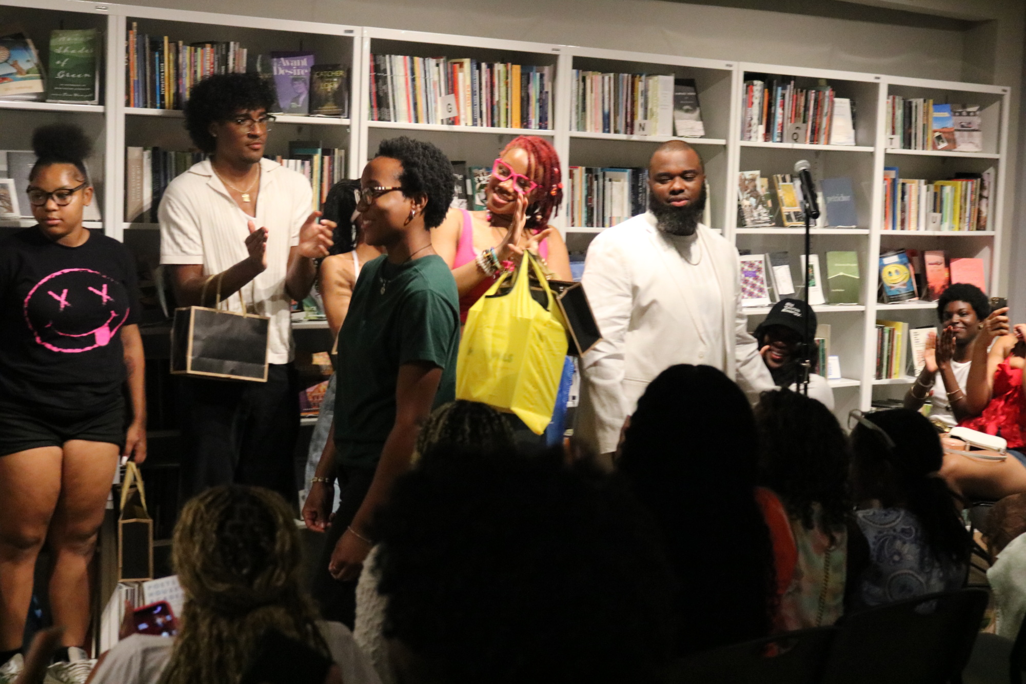 A diverse group of people is gathered in front of a bookshelf. Some hold bags and appear to listen intently, possibly during an event or presentation. The background shows many books and shelves.