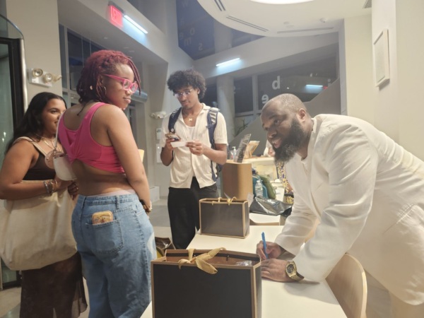 A man in a white suit signs a book at a table while three people stand nearby. Two of them watch him, and one person checks a phone.