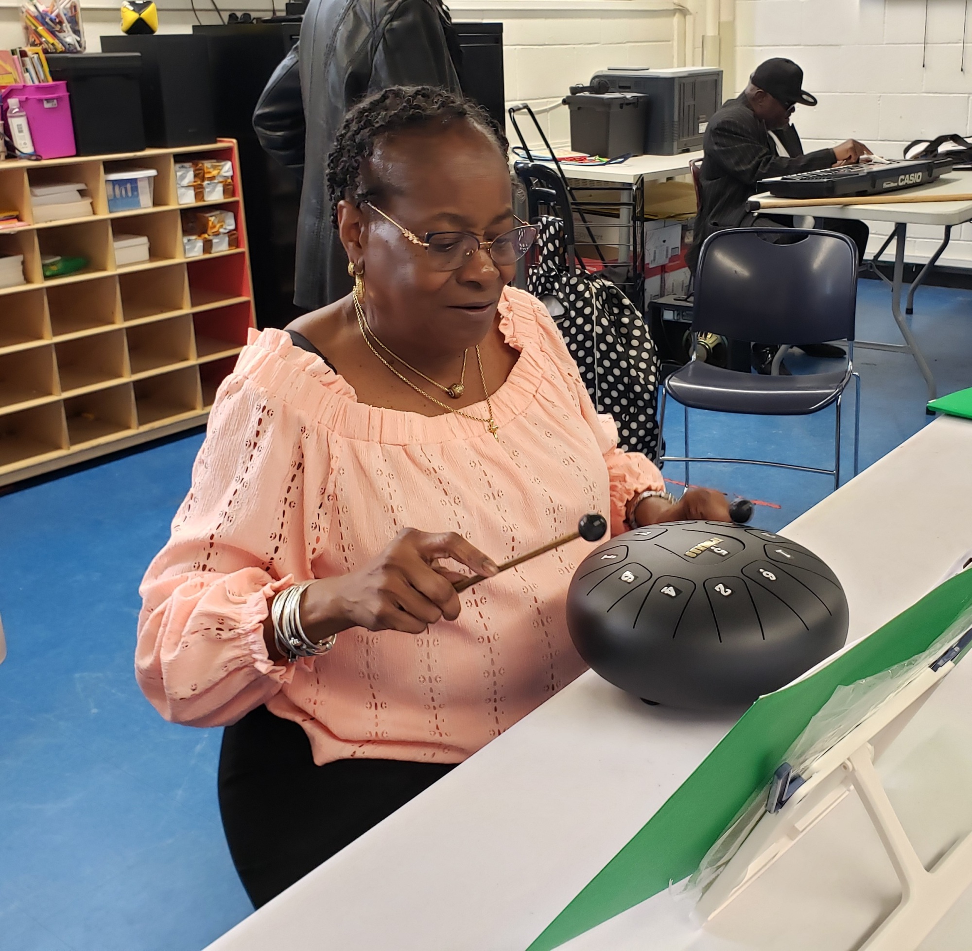 A woman plays a steel tongue drum with a mallet while sitting at a table in a classroom setting.