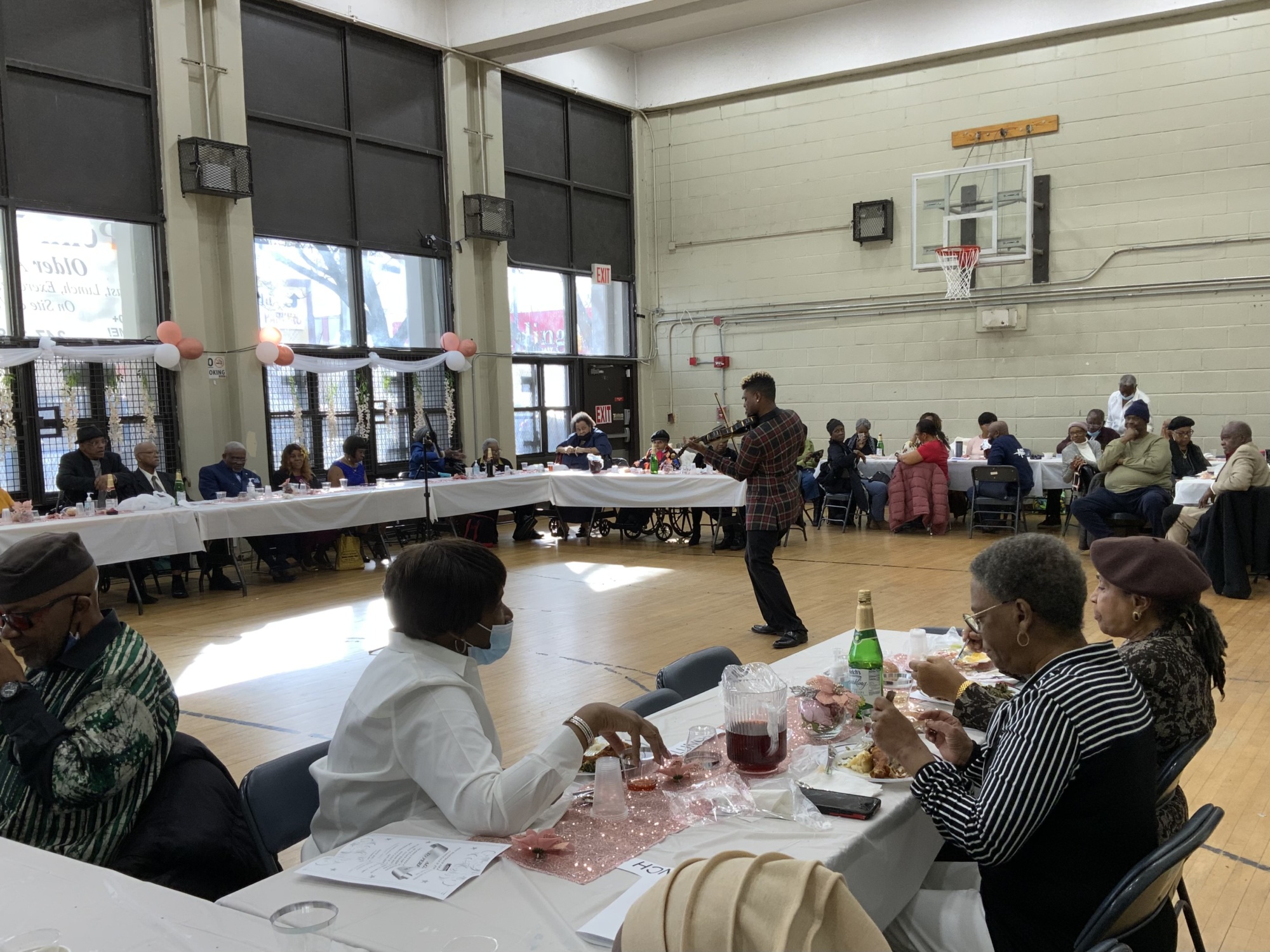 A person speaks into a microphone in the center of a gymnasium with tables arranged in a U-shape. Attendees are seated around the tables, eating, and listening. Pink balloons and decorations are visible.