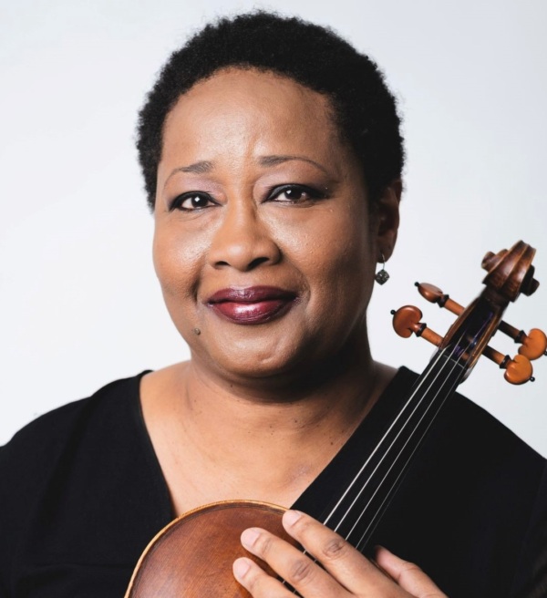 A person with short hair and a black outfit holds a violin, looking at the camera with a slight smile.