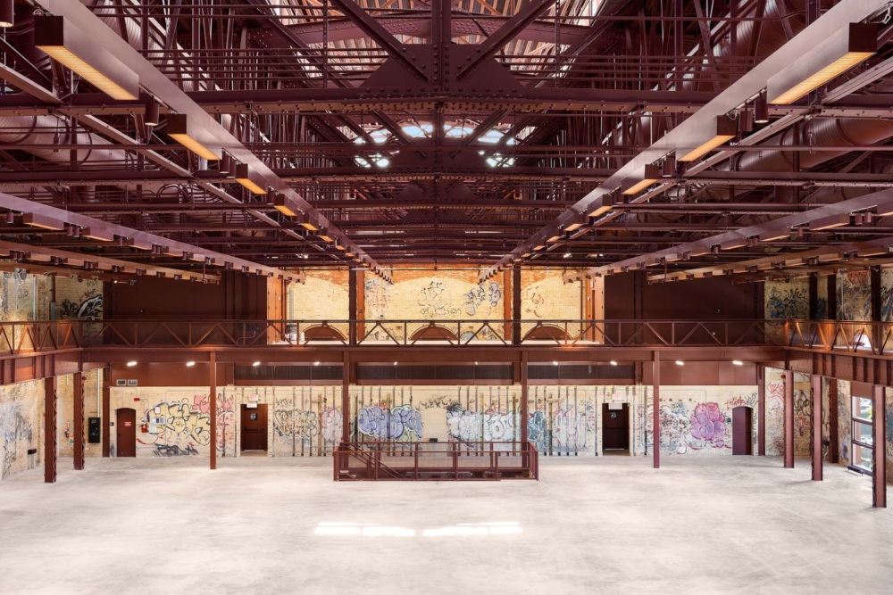 An industrial building with an open floor, high ceilings, brown steel beams, graffitied walls, and large windows allowing ample natural light.