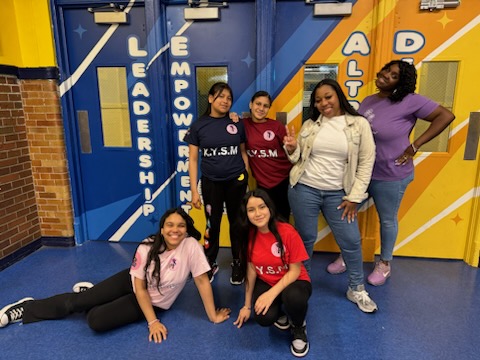 Six individuals pose and smile in front of doors labeled "Leadership," "Empowerment," and "Alphabet." They wear different colored t-shirts and appear to be in a school or community center.