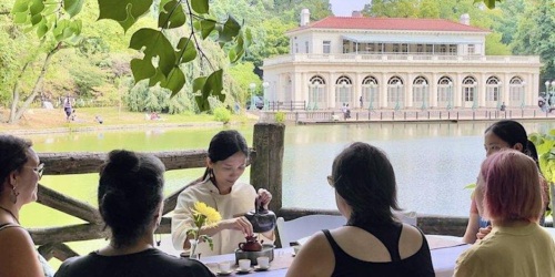 A group of people sit outside near a lake, engaging in a tea ceremony facilitated by one person. In the background, a large building with multiple arches is visible. Trees and greenery surround the scene.