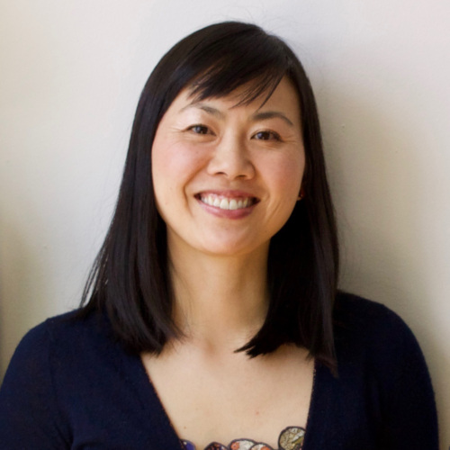 A woman with straight, dark hair smiles at the camera against a plain background while wearing a black top.