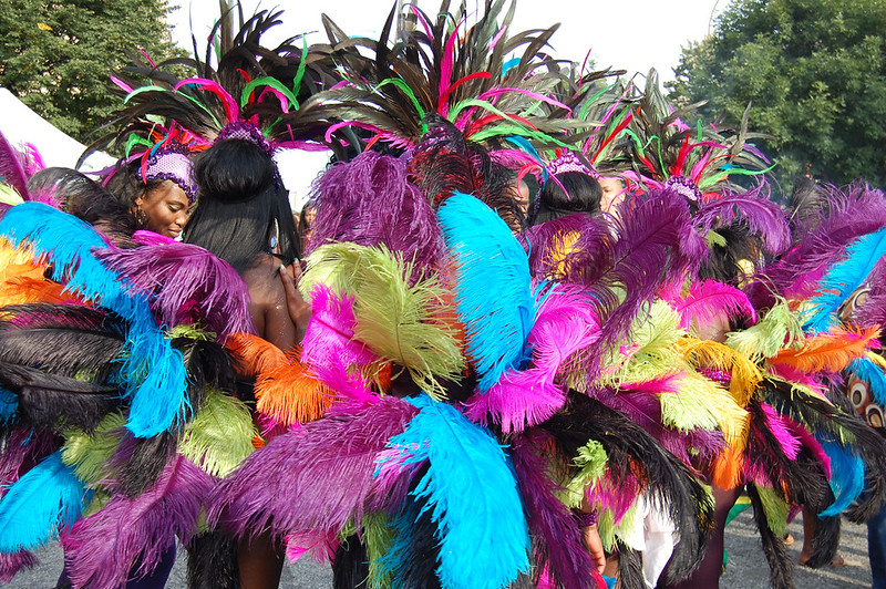 People wearing colorful feathered costumes with vibrant hues of purple, green, blue, pink, and orange. They are standing in a street, possibly at a festival or parade.