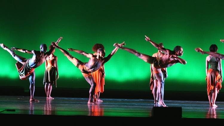 A group of dancers perform a synchronized routine on stage, each extending one leg high into the air, against a green lighted background. They're dressed in vibrant orange and green outfits.