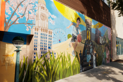 A colorful mural on a building wall depicts a boy playing basketball, Brooklyn landmarks, the Coney Island Wonder Wheel, and greenery under a sunny sky.