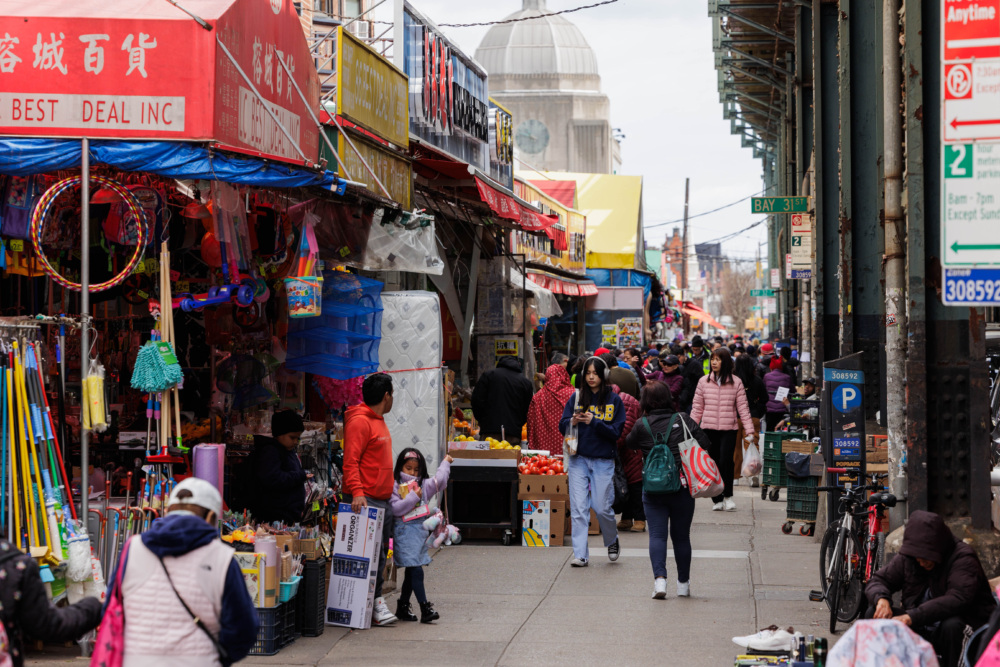 A busy street market with colorful stalls and signs, various merchandise on display, and numerous people walking and shopping.