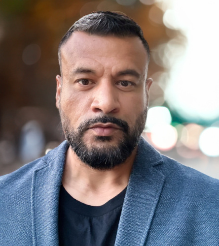 A man with a beard and short hair wearing a blue blazer and black shirt stands outdoors, with a blurred background of trees and lights.