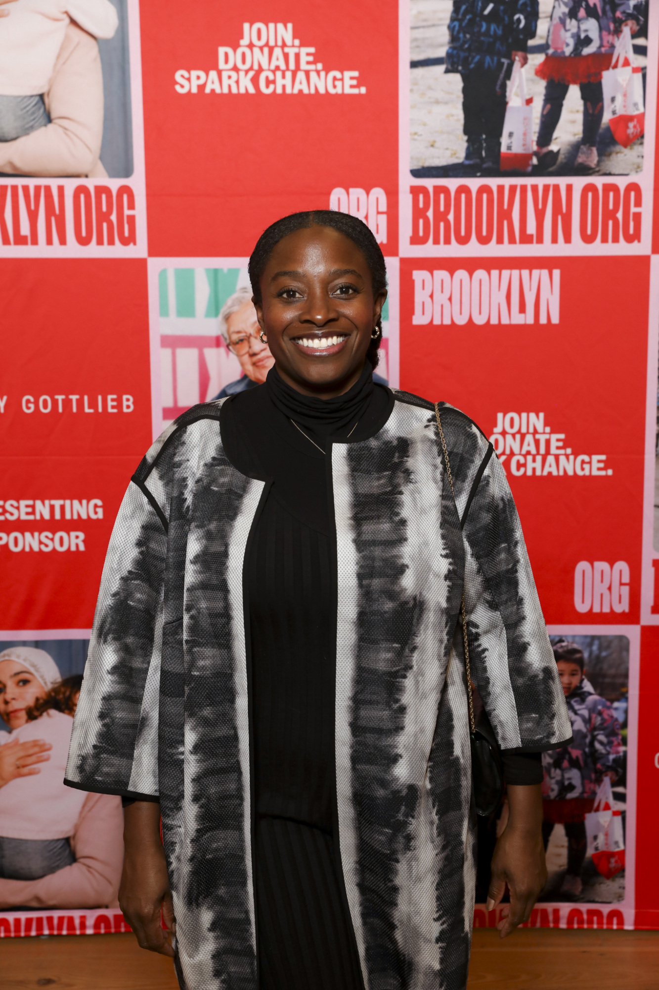 Woman smiling in front of a red and white poster wall with "Brooklyn" and "Join. Donate. Spark Change." text.