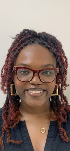 Smiling person with curly hair and large, square glasses, wearing hoop earrings and a necklace, against a plain background.