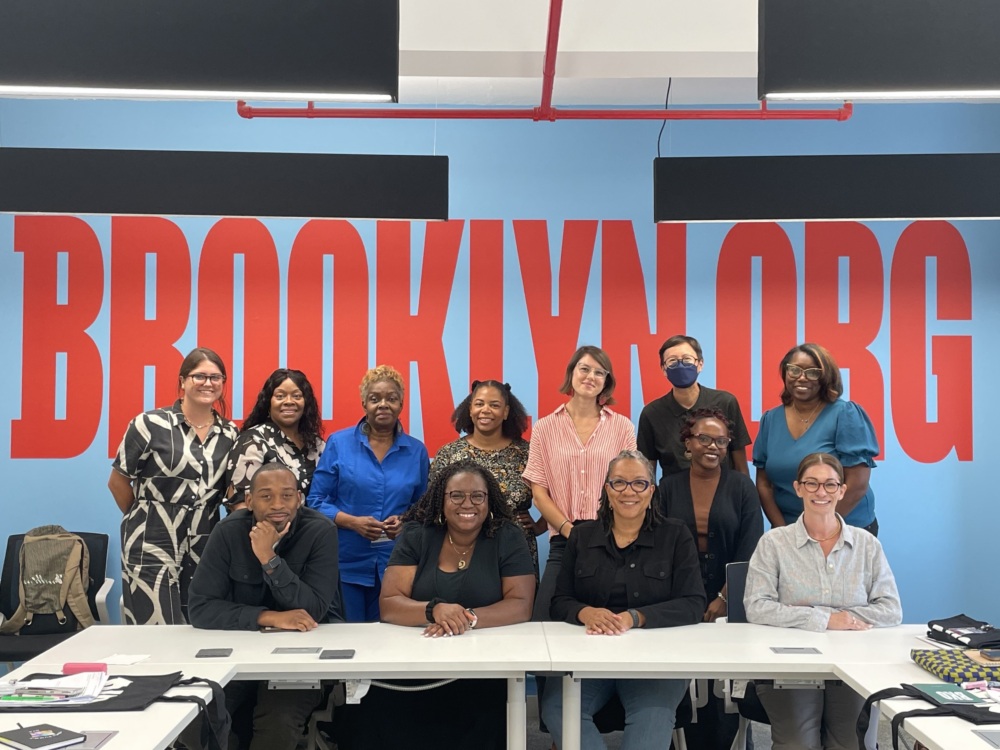 A group of people poses together in front of a large "BROOKLYN.ORG" sign, inside a room with blue walls and a white ceiling.