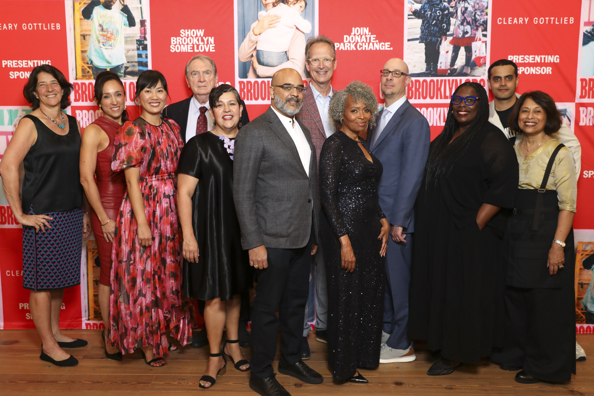 A group of twelve people stand in front of a red backdrop with text at an event. They are dressed in formal attire and pose together, smiling at the camera.