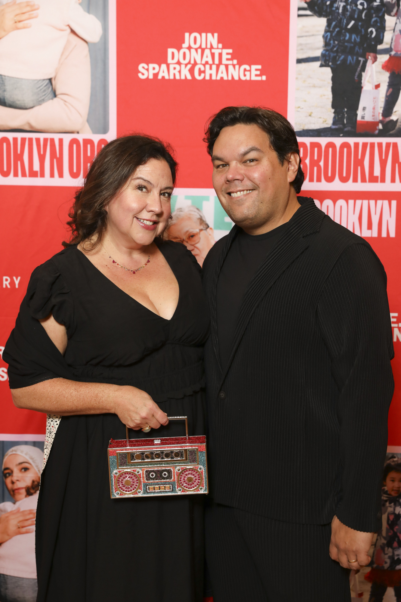 Two people in formal attire pose together, smiling in front of a red backdrop with the words "JOIN. DONATE. SPARK CHANGE.