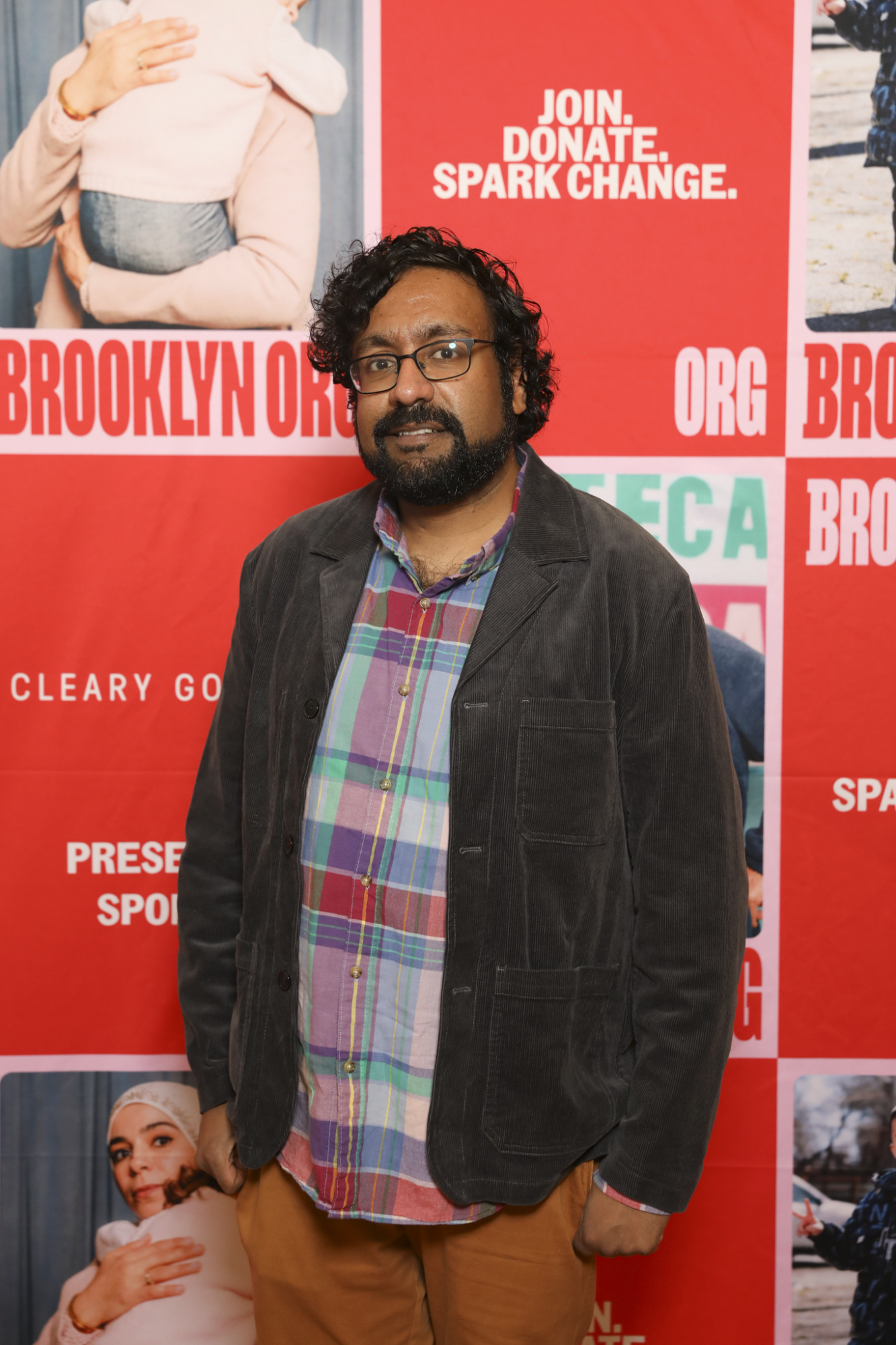 A person stands in front of a red and white backdrop featuring text "BROOKLYN ORG" and "JOIN. DONATE. SPARK CHANGE." They are wearing glasses and a plaid shirt under a corduroy jacket.