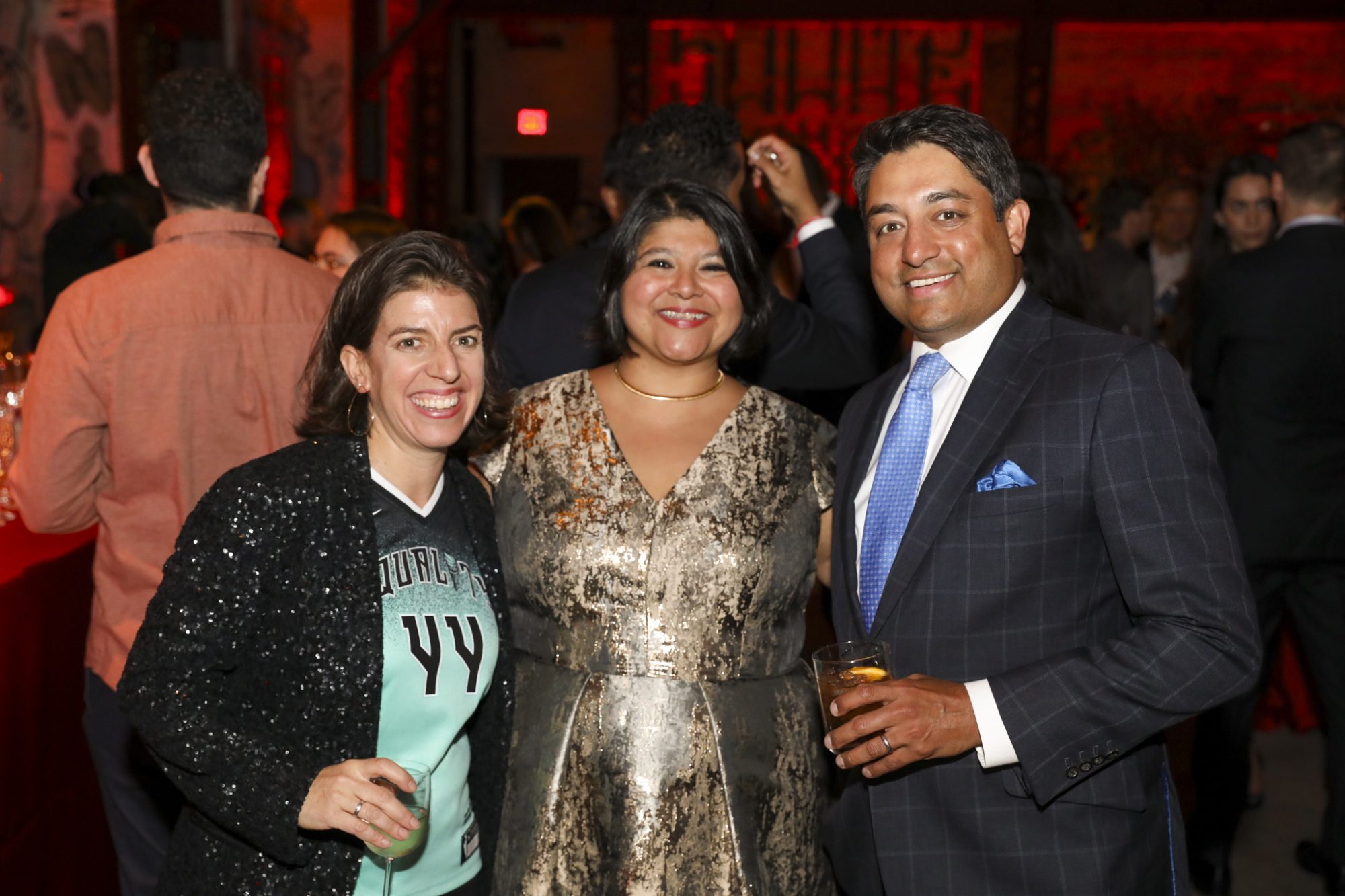 Three people smiling at an indoor event, with two holding drinks.