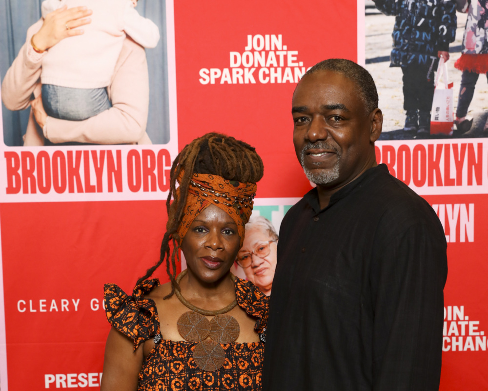 A man and woman stand together smiling in front of a Brooklyn Organics backdrop. The woman is wearing a patterned outfit and headwrap, while the man is dressed in a dark shirt.