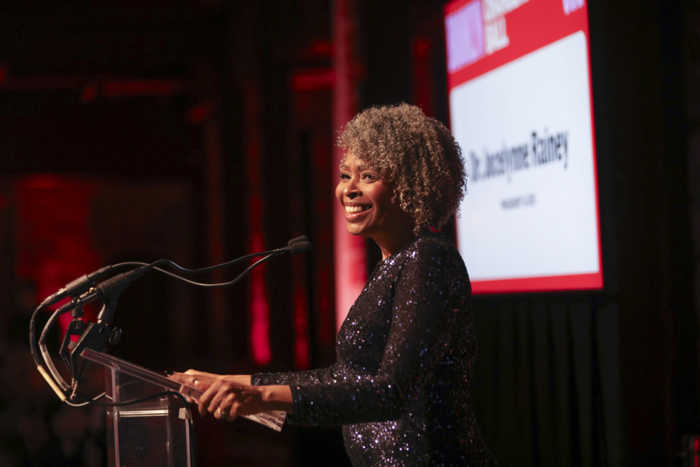 A smiling woman with curly hair speaks at a podium, with a screen displaying text in the background.