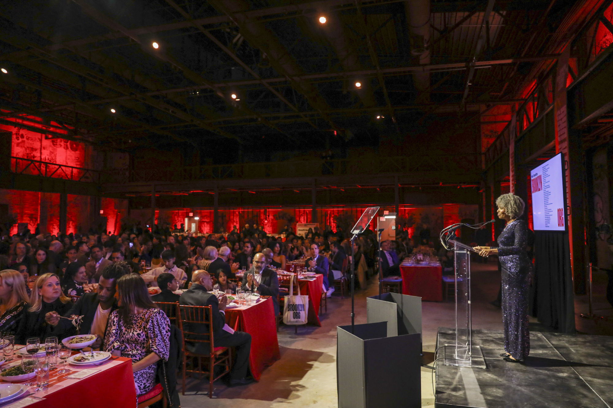 A woman speaks at a podium to a large seated audience in a dimly lit event space, with red ambient lighting.