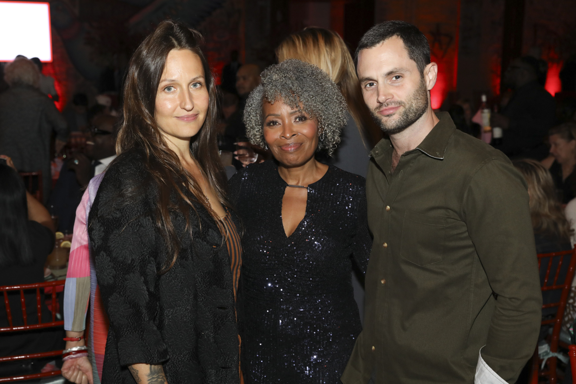 Three people standing together at an indoor event, with others in the background. The setting features tables and chairs, and the lighting has a warm tone.