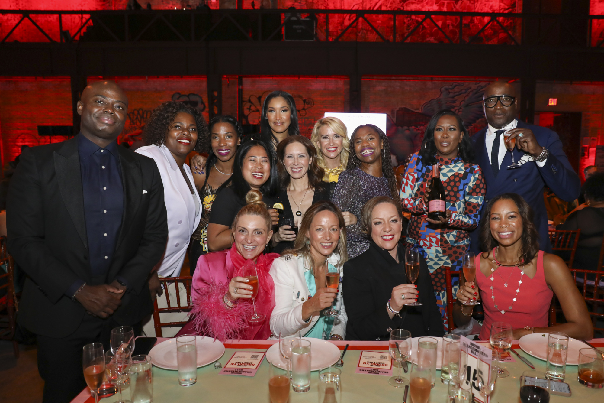 A group of people dressed formally, posing together at a dining table with drinks in hand, in an indoor event setting.