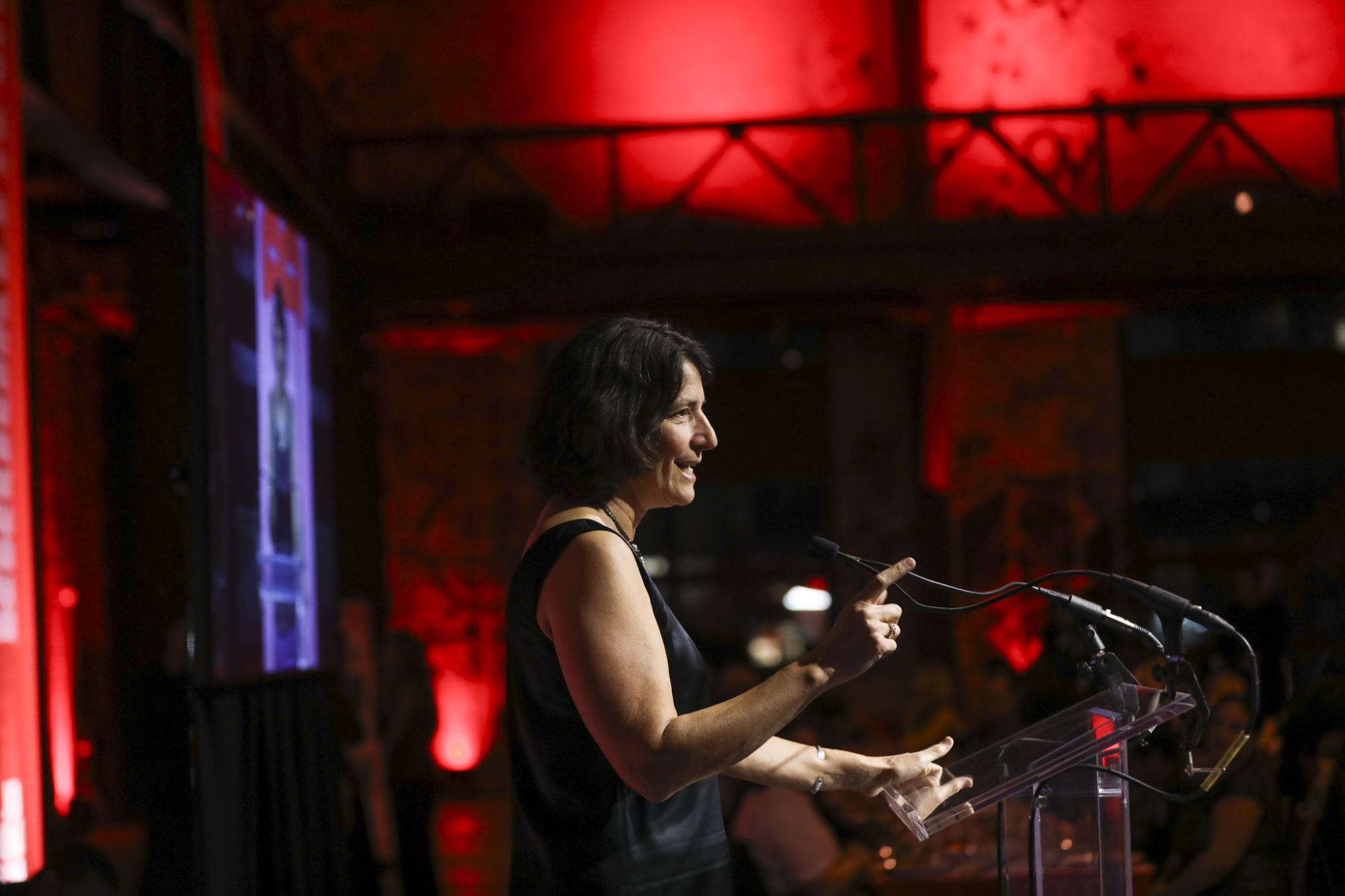 A person speaks at a podium with a microphone in a dimly lit room, with red lights in the background.