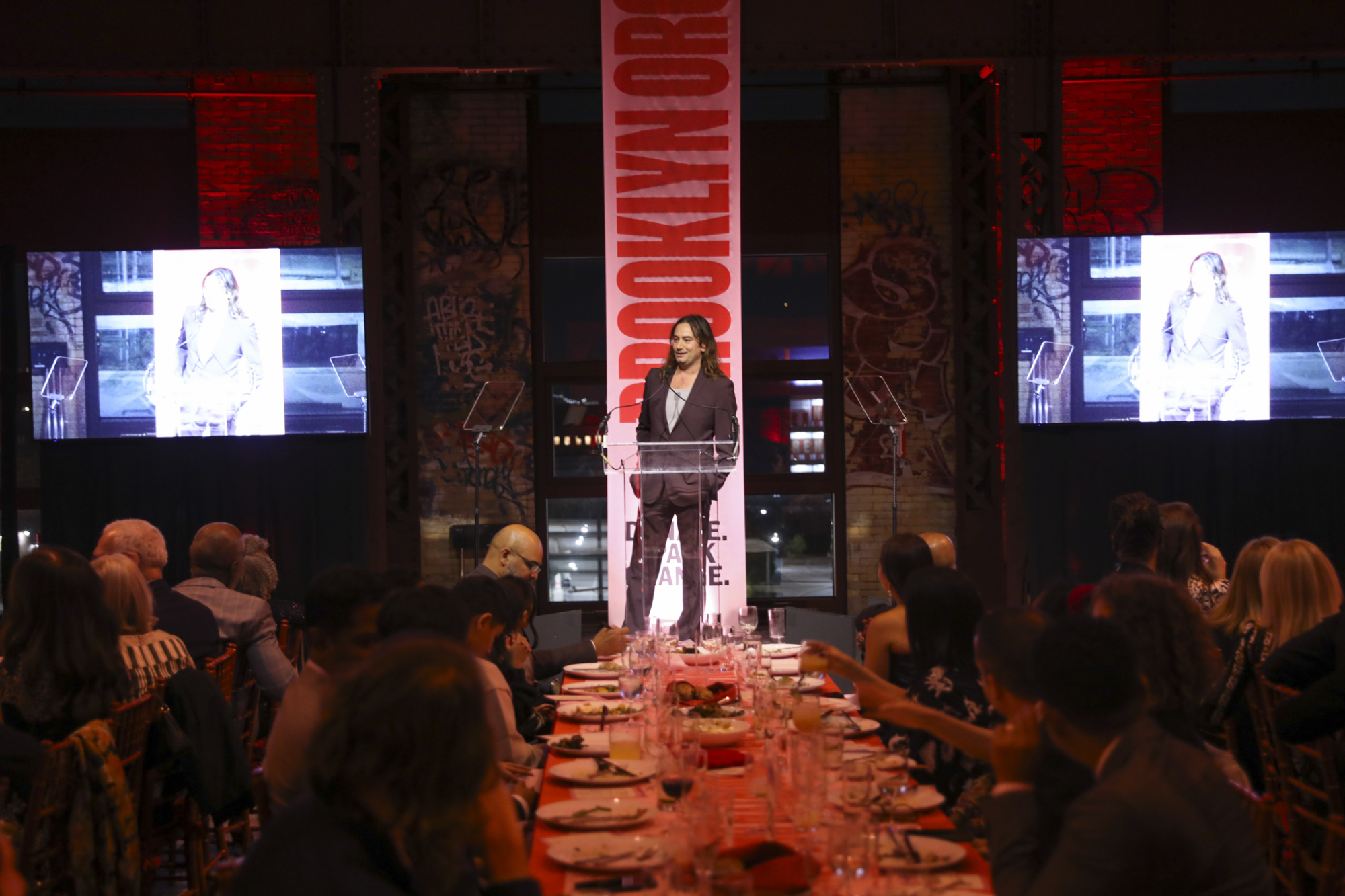 A person speaks at a podium on stage with two screens displaying the same image. An audience is seated at tables, and the backdrop includes "BROOKLYN ORG" text.