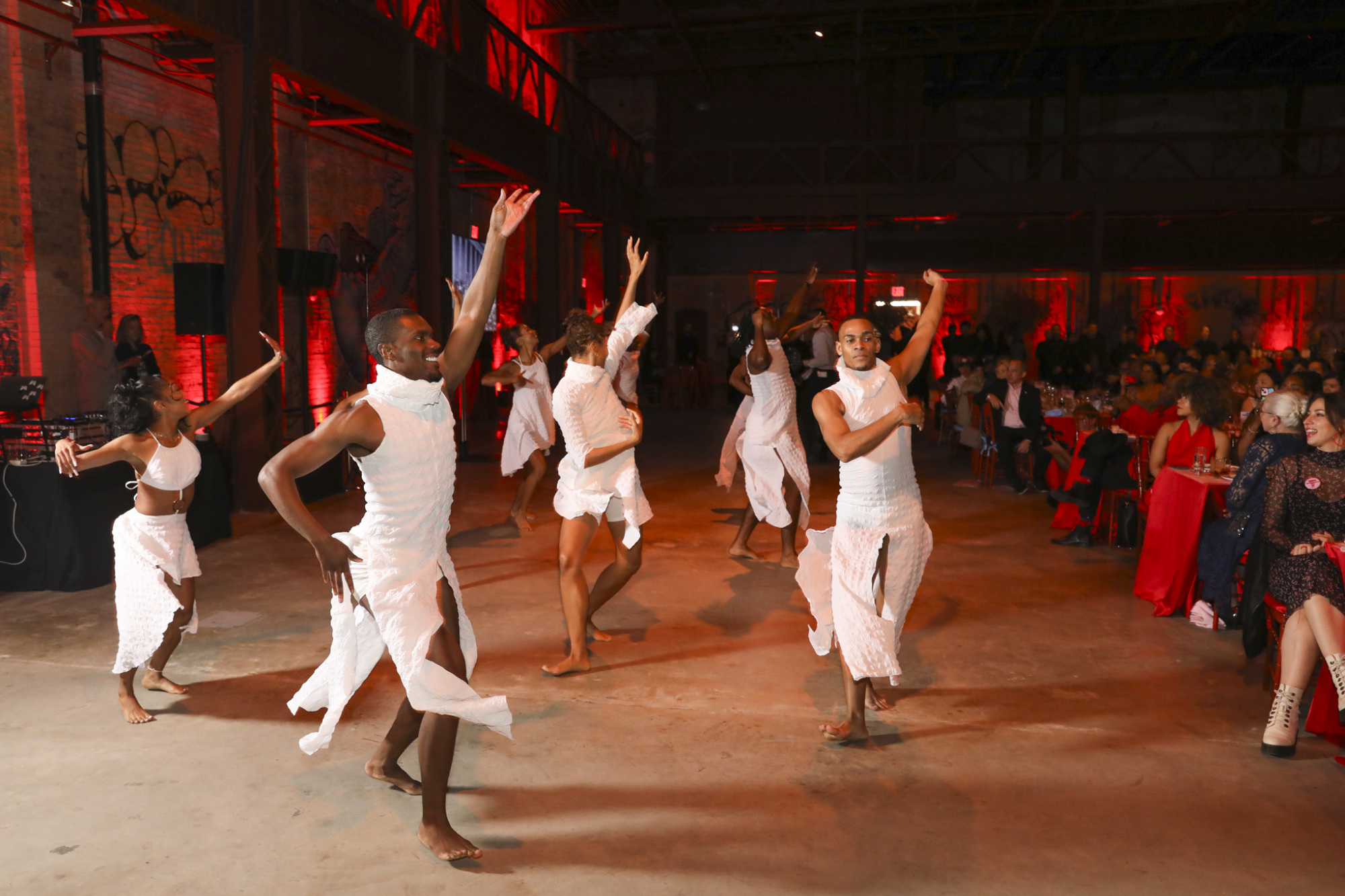 A group of six dancers in white costumes perform energetically on a stage in a dimly lit venue with red lighting, in front of a seated audience.