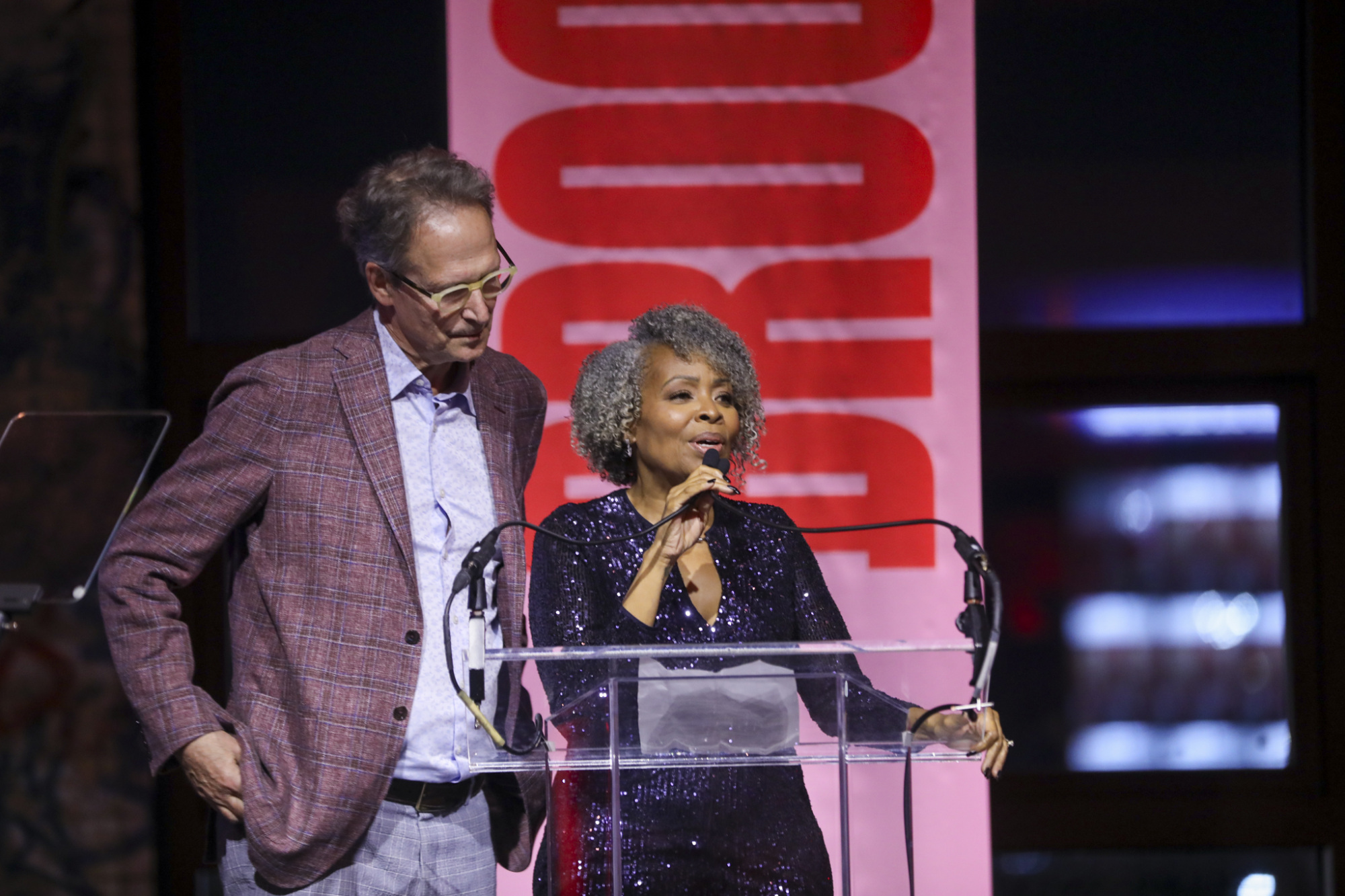 Two people stand at a podium. The woman speaks into a microphone, wearing a sparkly black outfit. The man, in a patterned jacket, stands beside her. A large red banner is in the background.