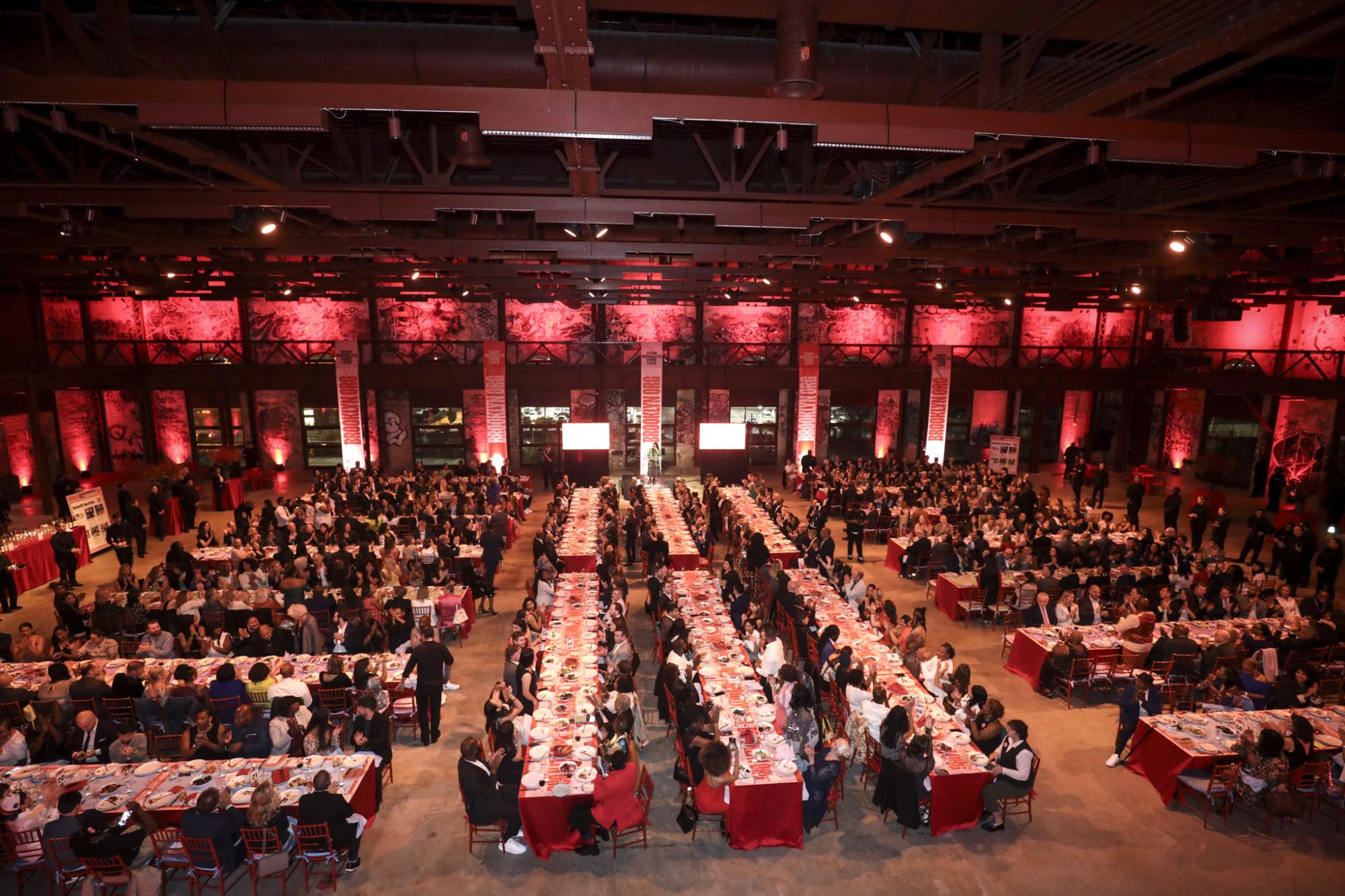 A large formal event with numerous attendees seated at long, parallel tables in a spacious venue lit with red lighting.