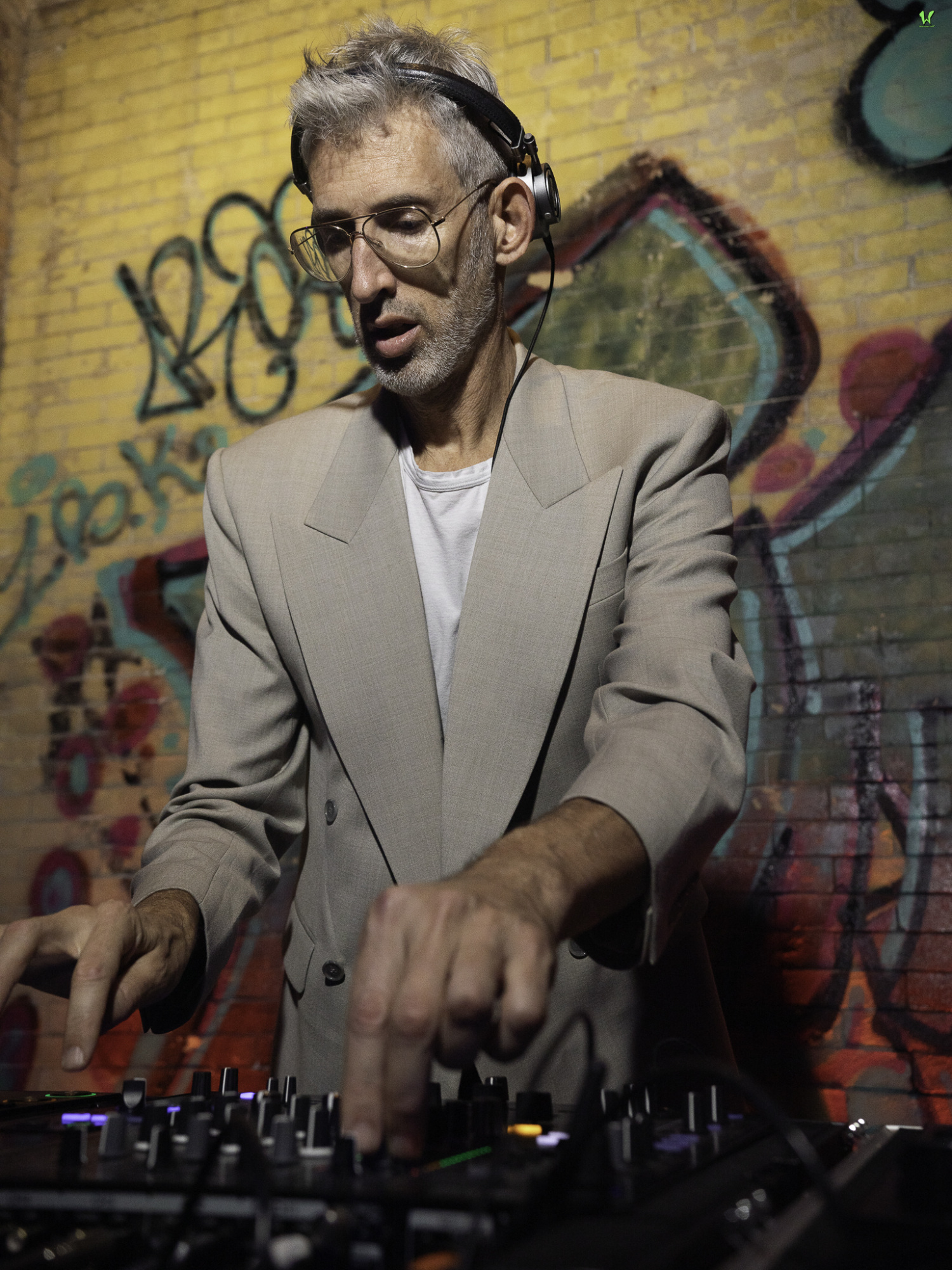 A man with headphones and glasses, wearing a beige blazer, is DJing in front of a graffiti-covered brick wall.