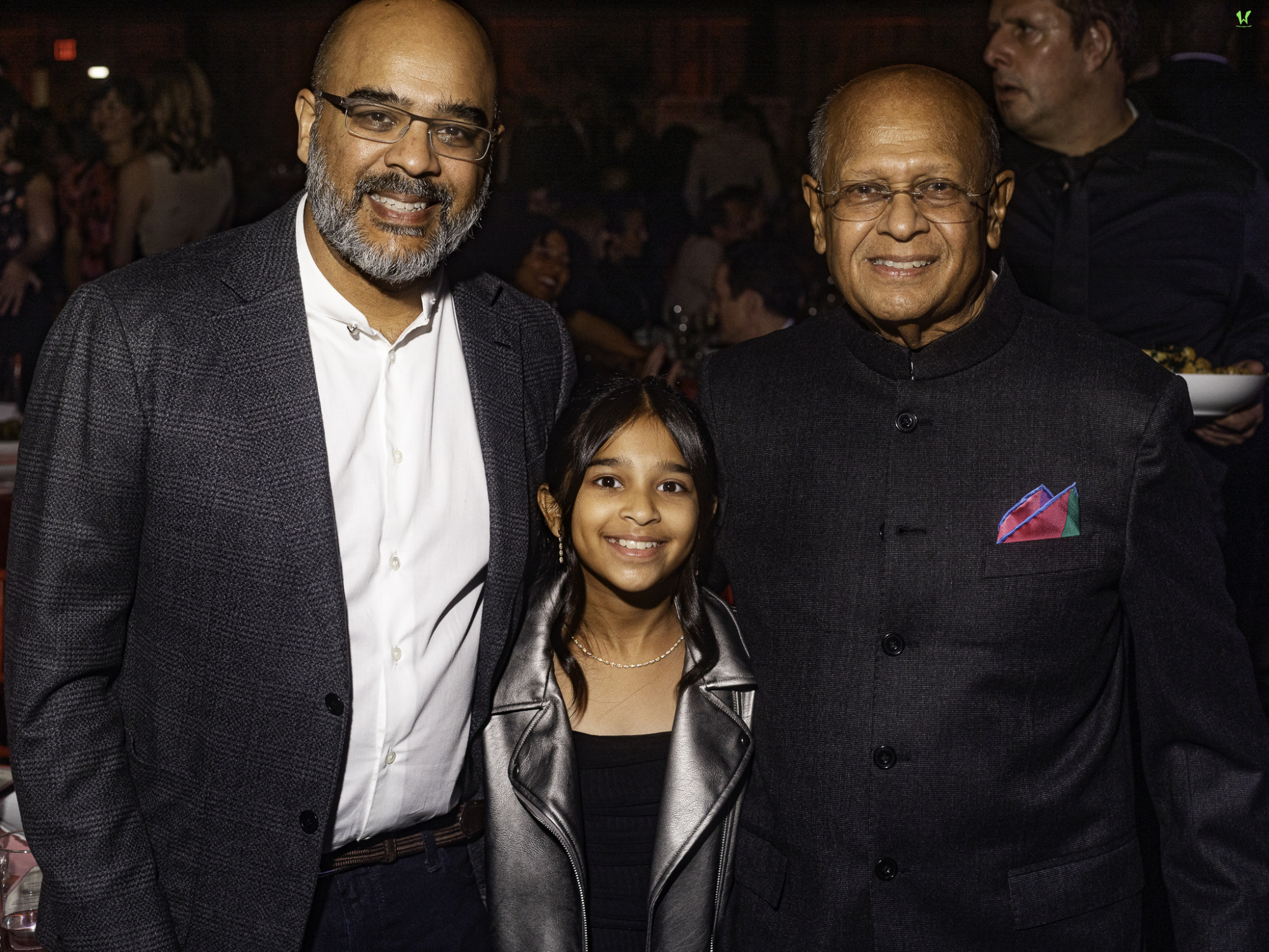 A young girl stands between two older men, all smiling at a social event. They are dressed in formal attire.