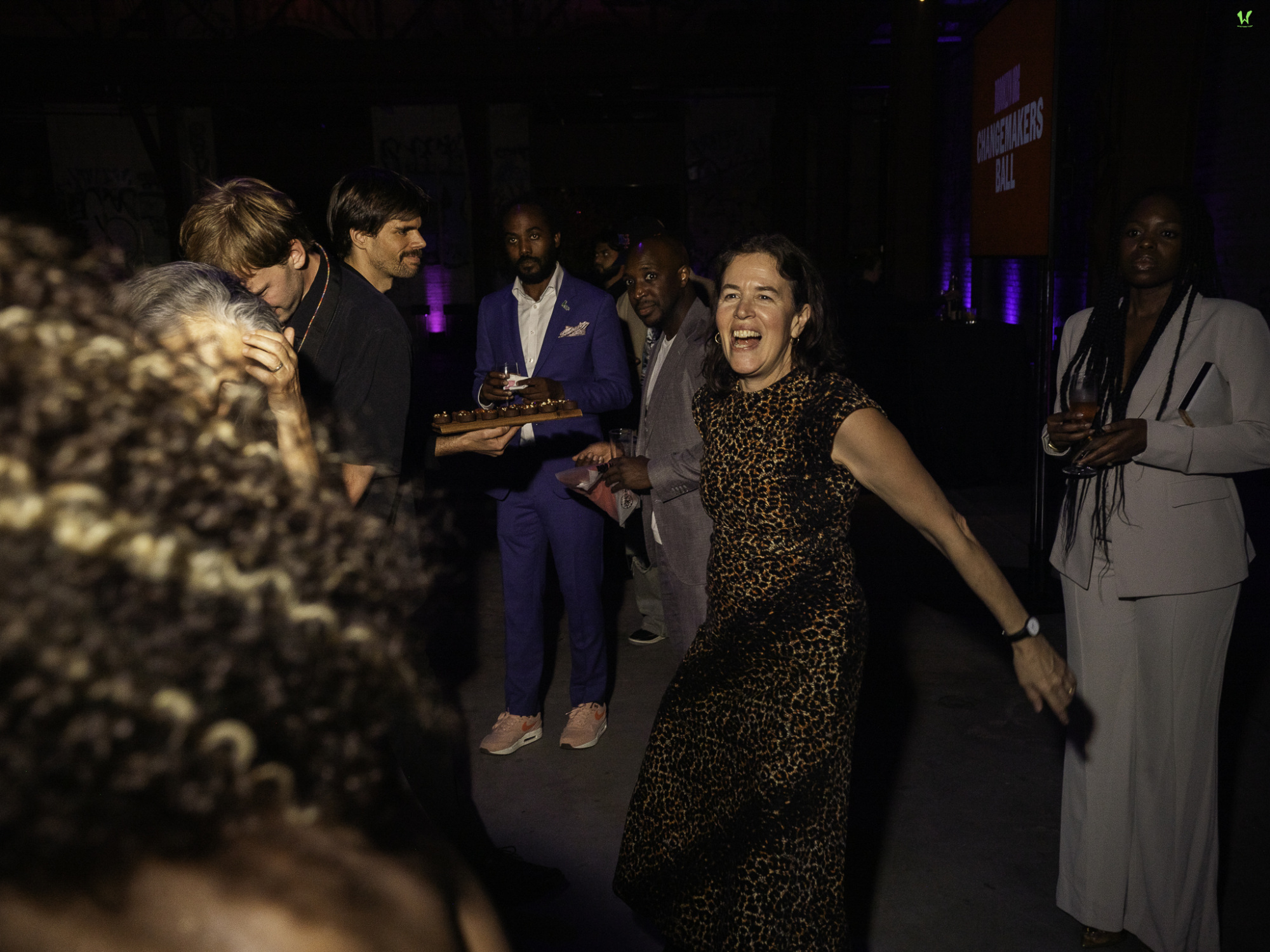 People socializing and dancing at an indoor event, with one woman in a leopard-print dress smiling in the foreground.