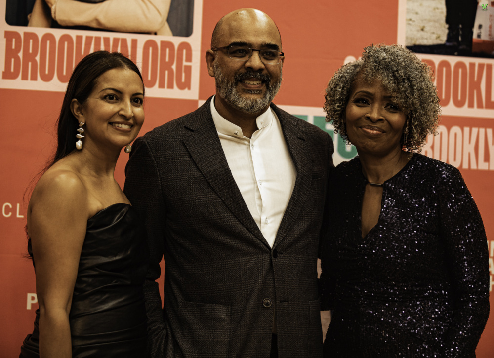 Three people pose together at an event, smiling in front of a backdrop with "Brooklyn" printed on it.