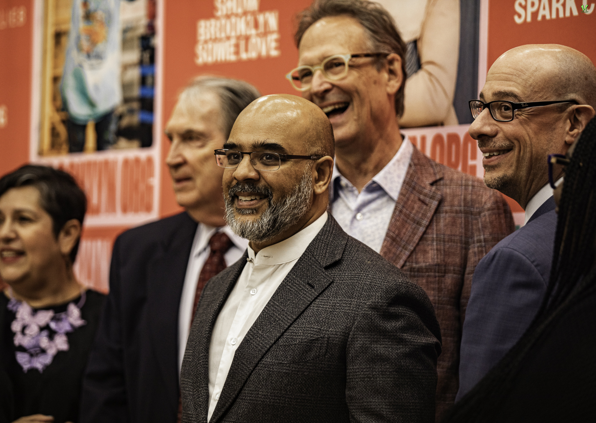 A group of six people in formal attire standing in front of a red backdrop with text. They are smiling and looking in various directions.