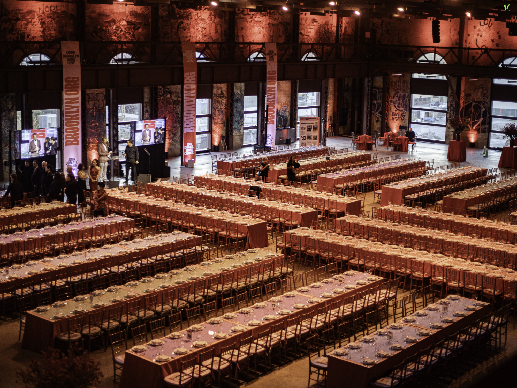 Large banquet hall with rows of long tables set for an event. Warm lighting and decorative posters are visible.