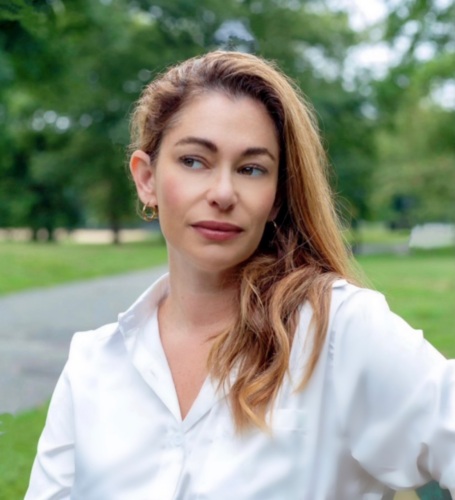 Woman with long hair in a white blouse stands outdoors in a park area with trees and grass in the background.