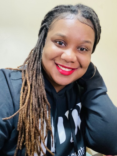 A person with long dreadlocks, wearing a black hoodie and hoop earrings, smiles while resting their head on their hand.