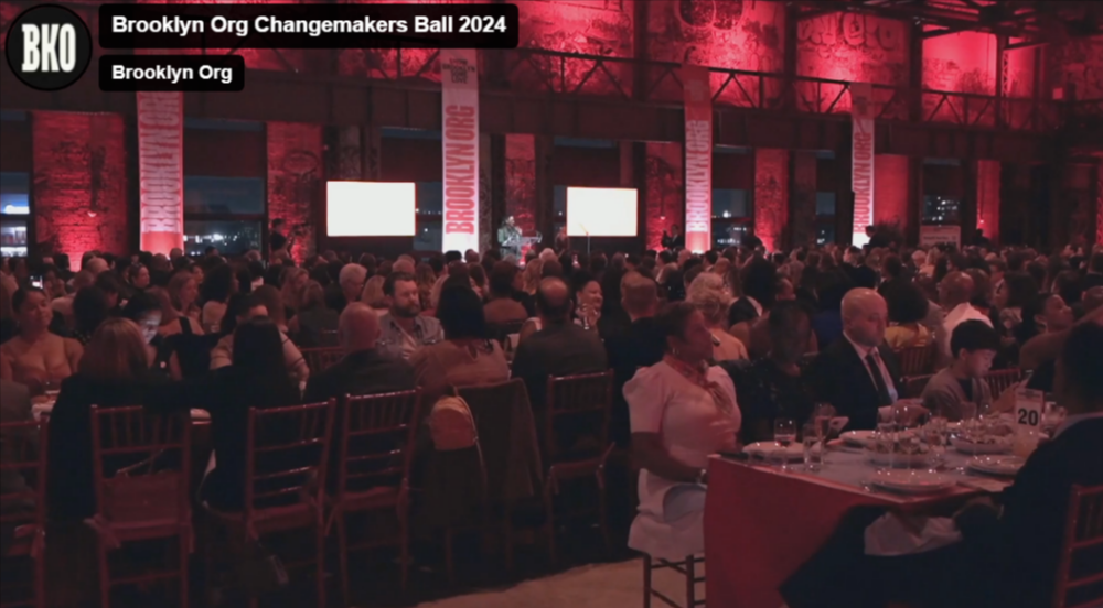 A large, indoor gala event with attendees seated at tables, red lighting, and the text "Brooklyn Org Changemakers Ball 2024" visible on banners.