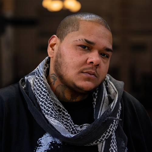 A person with a shaved head and facial hair, wearing a patterned scarf and a black shirt, looks directly at the camera with a serious expression.