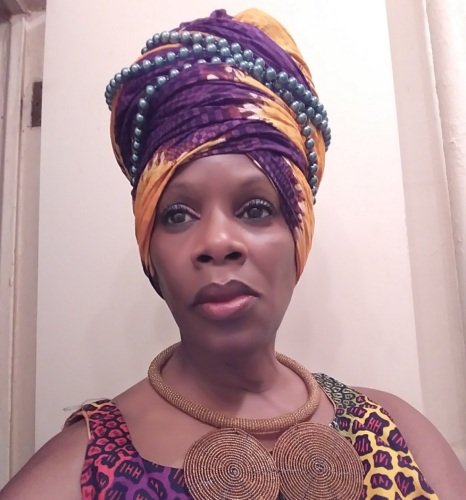 Person wearing a colorful headwrap and patterned outfit, adorned with large circular necklace, stands in front of a beige wall.