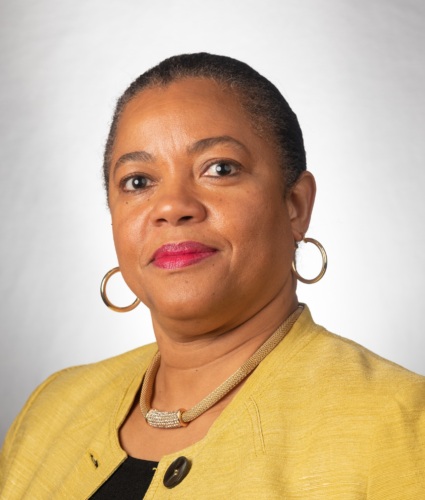 A person wearing a yellow jacket and hoop earrings stands against a neutral background.